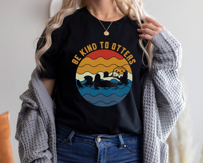 Pu&ograve; includere: Una maglietta nera con una grafica in stile vintage che dice "Sii gentile con le lontre". La grafica presenta un tramonto retr&ograve; con onde e una lontra di cartone animato.