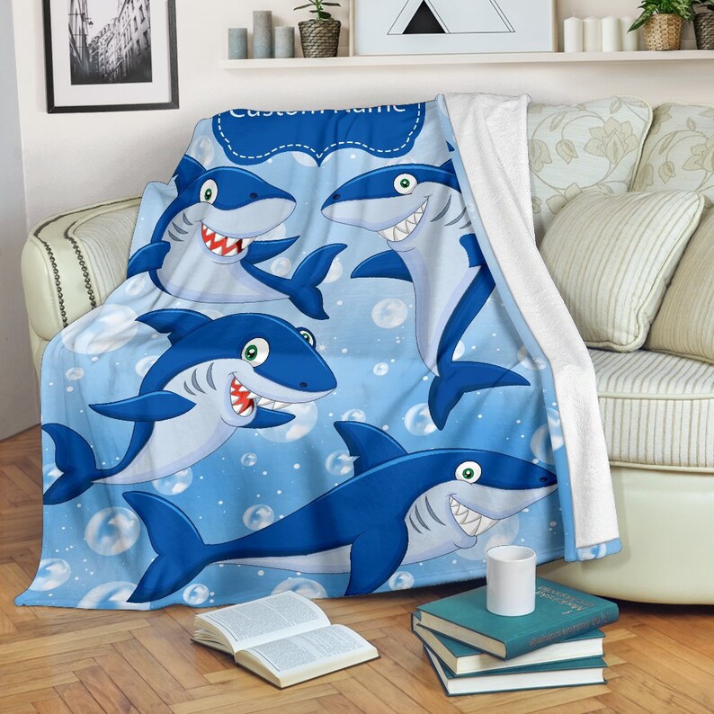 May include: A blue fleece throw blanket with a cartoon shark pattern. The blanket features multiple sharks with white teeth and blue bubbles on a light blue background. The text "Custom Name" is in a black banner at the top of the blanket.
