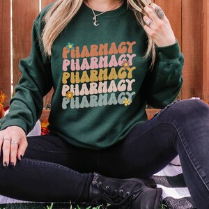 May include: A dark green sweatshirt with the word "Pharmacy" repeated five times in a retro font with colorful flowers. The sweatshirt is being worn by a person sitting on a striped blanket.