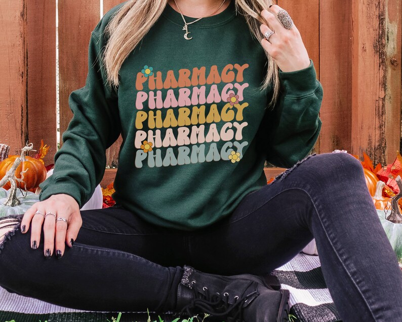 May include: A dark green sweatshirt with the word "Pharmacy" repeated five times in a retro font with colorful flowers. The sweatshirt is being worn by a person sitting on a striped blanket.