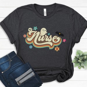 Op de afbeelding: Donkergrijs t-shirt met het woord "Nurse" in een retro lettertype, versierd met geesten, bloemen en een vleermuis. Het shirt wordt gecombineerd met een blauwe jeans, wat een casual, alledaagse look cre&euml;ert. Het ontwerp heeft een vintage, Halloween-ge&iuml;nspireerd thema.