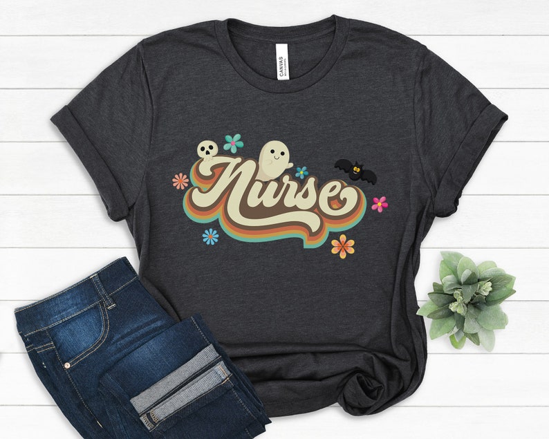 Op de afbeelding: Donkergrijs t-shirt met het woord "Nurse" in een retro lettertype, versierd met geesten, bloemen en een vleermuis. Het shirt wordt gecombineerd met een blauwe jeans, wat een casual, alledaagse look cre&euml;ert. Het ontwerp heeft een vintage, Halloween-ge&iuml;nspireerd thema.