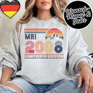 May include: A light gray sweatshirt with a retro design featuring the word "MAI" above the number "2008" in a colorful, striped font. The design includes a mountain graphic and the words "LIMITED EDITION". A black circle with white text is in the upper right.