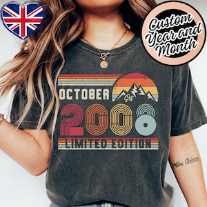 May include: A charcoal gray t-shirt featuring a retro-style graphic with the text "OCTOBER", "EST", "2008", and "LIMITED EDITION". The design includes a sunset over mountain peaks in shades of orange, yellow, and blue, with concentric line patterns.