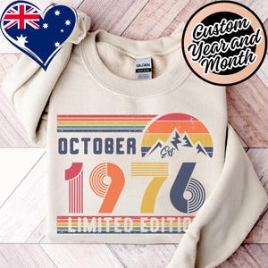 May include: A cream-coloured crewneck sweatshirt featuring a graphic print with the text OCTOBER 1976 LIMITED EDITION. The design includes a retro-style sunset, mountain silhouettes, and horizontal stripes in shades of red, orange, yellow, and blue.