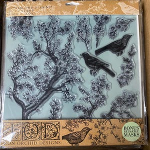 May include: A set of clear rubber stamps featuring a floral design with birds. The stamps are packaged in a clear plastic sleeve with a brown and green background. The text "Iron Orchid Designs" and "Bonus Reusable Masks" is visible on the packaging.