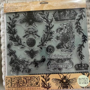 May include: Clear stamp set featuring bee and crown designs. Includes text "IRON ORCHID DESIGNS", "QUEEN BEE", and "JEAN FRANÇOIS-GILLET, ECUYER, 1778". Contains bonus reusable masks. Ideal for crafting and scrapbooking projects.