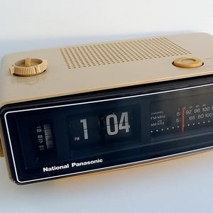 Może przedstawiać: Beżowy radiobudzik z klapkami National Panasonic z czarnym panelem przednim. Urządzenie posiada cyfrowy zegar z klapkami wskazujący godzinę 1:04 PM, analogowy tuner radiowy z częstotliwościami FM i AM, kratkę głośnika oraz okrągłe pokrętła sterujące.