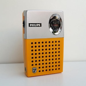 May include: A retro Philips radio in silver and yellow. The radio has a clock and the Philips logo. The front is perforated with small holes. A vintage electronic device.