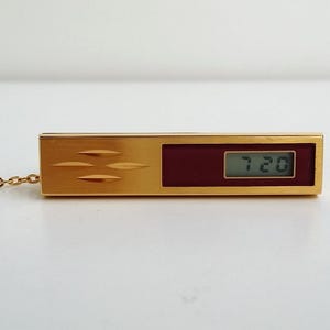 May include: A gold-toned rectangular pendant necklace with a digital clock display showing "7:20". The pendant has a burgundy-colored section and a chain. The pendant has three horizontal etched lines.