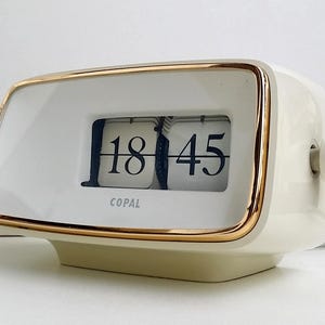 Könnte beinhalten: Eine cremefarbene Copal-Klappzahlenuhr mit goldfarbenem Metallrand um das Display. Die Uhr verfügt über eine digitale Klappanzeige, die die Uhrzeit 18:45 in schwarzen Ziffern auf weißen Plättchen anzeigt. Ein Einstellknopf befindet sich rechts.