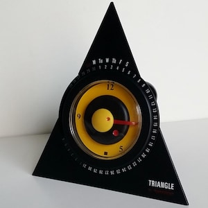 Op de afbeelding: Een zwarte driehoekige klok met een geel en zwarte cirkelvormige wijzerplaat. De klok heeft een rode secondewijzer en een zwarte driehoekige basis met een kalender. Het woord "TRIANGLE" staat op de basis.