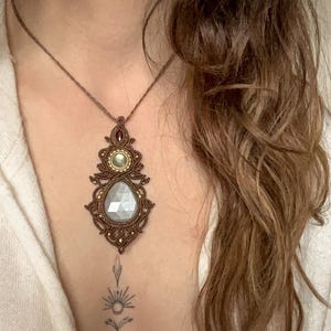 Op de afbeelding: Een handgemaakte macramé ketting met een grote, peervormige, lichtgrijze edelsteen hanger. De hanger is omlijst door ingewikkelde bruine macramé details, met gouden, groene en rode accenten. De ketting wordt gedragen aan een bruin koord.