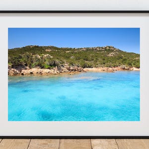 Könnte beinhalten: Gerahmtes Foto einer Küstenlandschaft. Das Bild zeigt klares türkisfarbenes Wasser, einen Sandstrand und einen Hügel, der mit grüner Vegetation bedeckt ist, unter einem strahlend blauen Himmel. Das Foto wird in einem schwarzen Rahmen präsentiert.