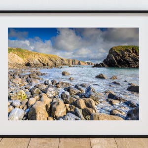 Op de afbeelding: Een ingelijste foto van een kustlandschap. De afbeelding toont een strand met veel grote, afgeronde stenen op de voorgrond. Het water is turquoise en de lucht is blauw met dramatische wolken. De kliffen zijn zichtbaar op de achtergrond.