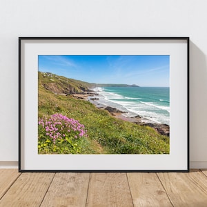 May include: A framed print of a coastal landscape. The image features a vibrant blue sky, turquoise ocean, and a shoreline with waves. Lush green vegetation and pink flowers are in the foreground. The print is in a black frame and rests on a wooden surface.