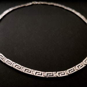 May include: A silver necklace with a Greek key pattern. The necklace is made of a thin chain and has a simple design.