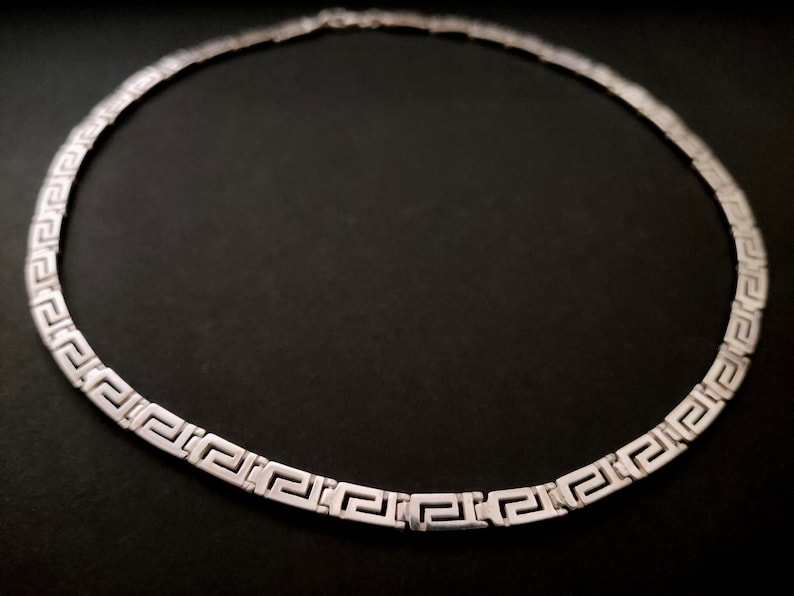 May include: A silver necklace with a Greek key pattern. The necklace is made of a thin chain and has a simple design.