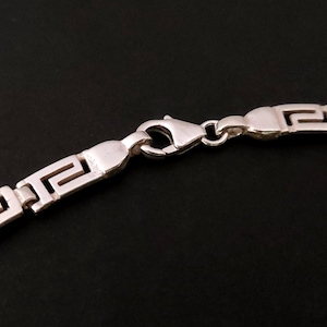 May include: Silver chain necklace with a Greek key pattern and a lobster clasp closure.