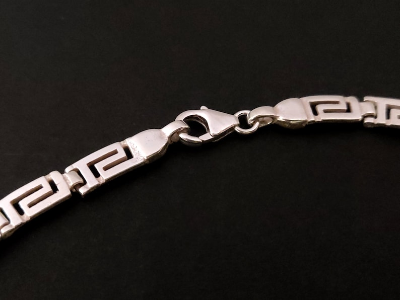 May include: Silver chain necklace with a Greek key pattern and a lobster clasp closure.