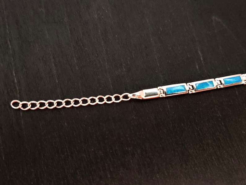 Puede incluir: Un brazalete de plata con una cadena y piedras rectangulares azules.