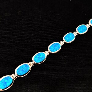 May include: A silver bracelet featuring oval-shaped, bright blue opal stones. The stones are set in a silver link chain. The bracelet is displayed against a black background, highlighting the vibrant blue color of the opals.