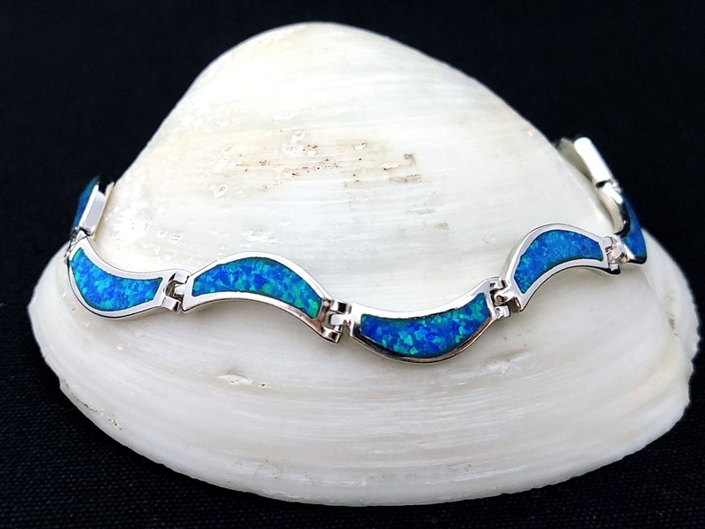 May include: A silver bracelet with a wave-like design. The bracelet features vibrant blue opal stones set in a polished silver frame. The opal stones have a mix of blue and green hues. The bracelet is displayed on a white seashell.