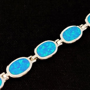 May include: A silver bracelet featuring oval-shaped blue opal stones. The opals have a vibrant, iridescent quality, with shades of blue and green. The bracelet is set against a black background.