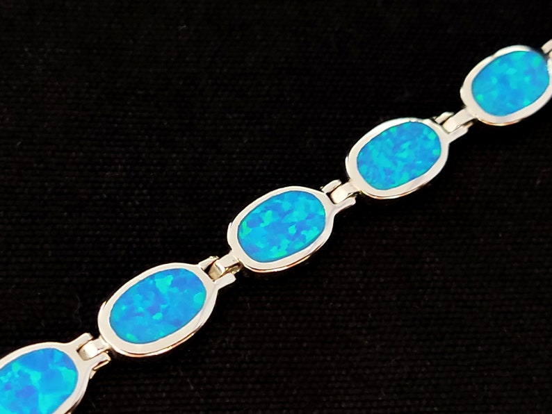 May include: A silver bracelet featuring oval-shaped blue opal stones. The opals have a vibrant, iridescent quality, with shades of blue and green. The bracelet is set against a black background.
