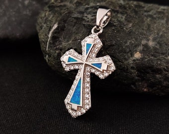 May include: A silver pendant in the shape of a cross, featuring a central blue opal design and surrounded by sparkling clear crystals. The cross is set against a dark, textured background.