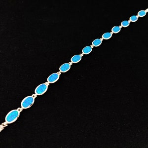 May include: A silver bracelet featuring a series of oval-shaped, bright blue opal stones. The stones are set in a silver chain, creating a delicate and elegant piece of jewelry. The bracelet is displayed against a black background.
