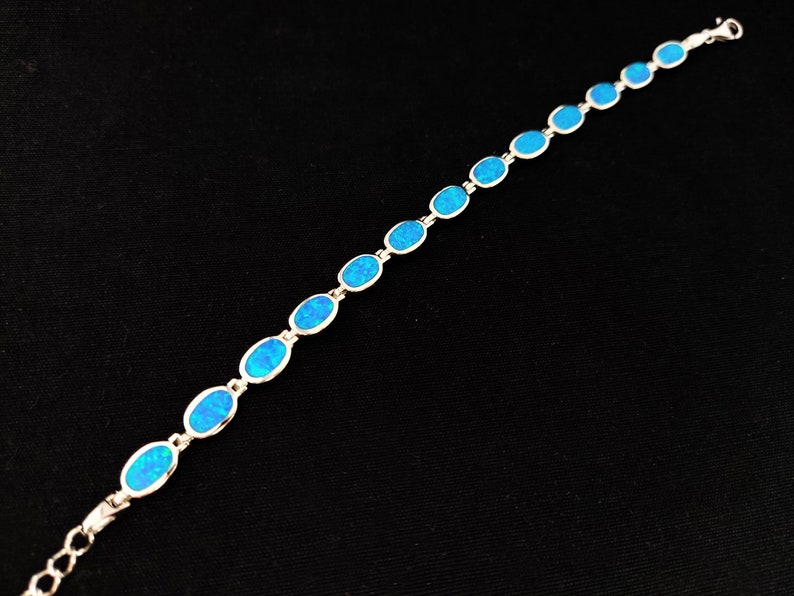 May include: A silver bracelet featuring a series of oval-shaped, bright blue opal stones. The stones are set in a silver chain, creating a delicate and elegant piece of jewelry. The bracelet is displayed against a black background.