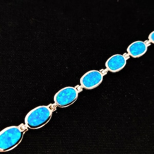May include: A silver bracelet featuring oval-shaped, bright blue opal stones. The stones are set in a silver link chain. The bracelet is displayed against a black background, highlighting the vibrant blue color of the opals.