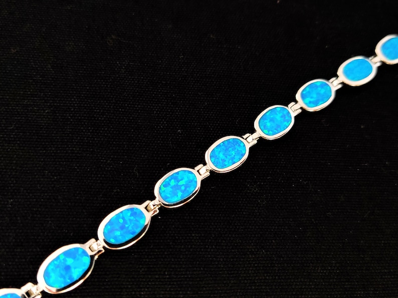May include: A silver bracelet featuring oval-shaped, bright blue opal stones. The stones are set in a silver link chain. The bracelet is displayed against a black background, highlighting the vibrant blue color of the opals.