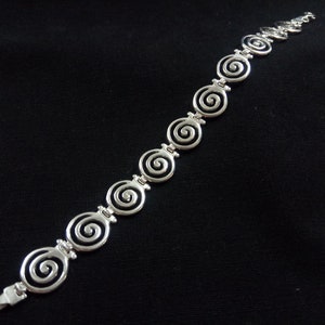 May include: Silver-toned bracelet featuring a series of spiral designs. The bracelet is composed of interconnected spiral links, creating a continuous pattern. The clasp is visible at the end of the bracelet. The jewelry is set against a black background.