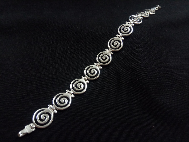 May include: Silver-toned bracelet featuring a series of spiral designs. The bracelet is composed of interconnected spiral links, creating a continuous pattern. The clasp is visible at the end of the bracelet. The jewelry is set against a black background.