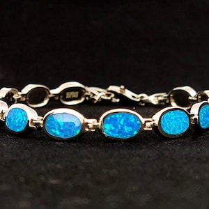 以下が含まれることがあります： 楕円形の鮮やかなブルーオパールストーンが特徴のシルバーブレスレット。 ブレスレットは繊細なチェーンデザインで、各オパールストーンはシルバーベゼルにセットされています。 クラスプもシルバーです。 黒い背景に表示されています。
