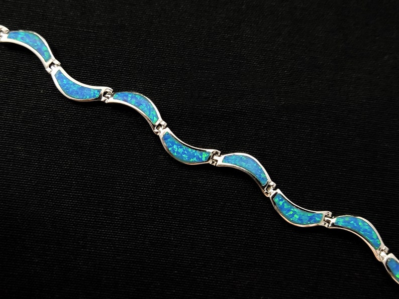May include: A silver bracelet with a wave-like design. Each wave is inlaid with a vibrant blue opal, creating a shimmering effect. The bracelet is composed of several connected segments.