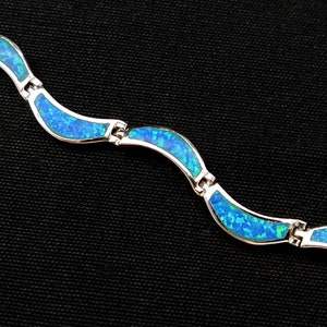 May include: A silver bracelet with a wave-like design. Each wave is filled with vibrant blue opal, creating a striking contrast against the silver. The bracelet is displayed against a black background.