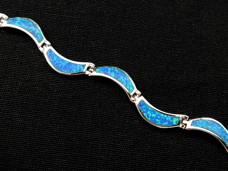 May include: A silver bracelet with a wave-like design. Each wave is filled with vibrant blue opal, creating a striking contrast against the silver. The bracelet is displayed against a black background.