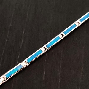 Op de afbeelding: Een zilveren armband met rechthoekige blauwe opaal inleg. De armband bestaat uit onderling verbonden schakels, elk met een levendige blauwe opaalsteen. Het ontwerp is eenvoudig en elegant.