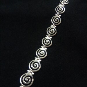 May include: A silver bracelet featuring a series of spiral designs. The bracelet is made of a shiny metal and is displayed against a black background. The spirals are evenly spaced and connected by small links.