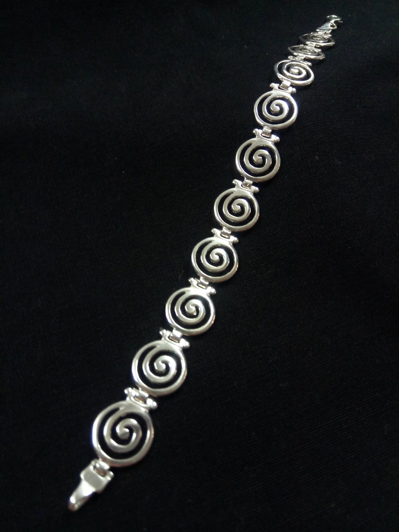 May include: A silver bracelet featuring a series of spiral designs. The bracelet is made of a shiny metal and is displayed against a black background. The spirals are evenly spaced and connected by small links.