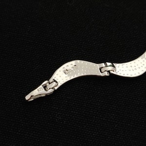May include: Close-up of a silver necklace clasp against a black background. The necklace has a textured, hammered design. The clasp is connected to a curved section of the necklace. The metal is stamped with "925".
