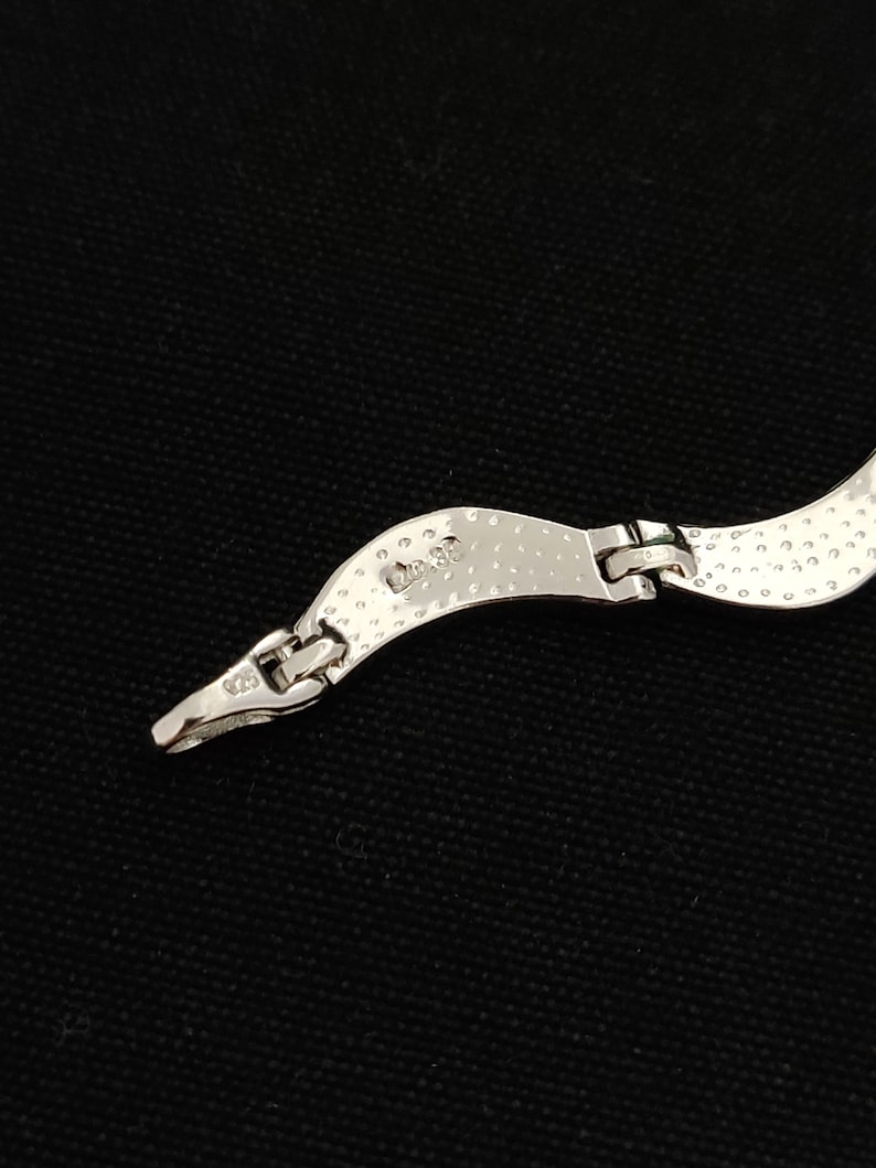 May include: Close-up of a silver necklace clasp against a black background. The necklace has a textured, hammered design. The clasp is connected to a curved section of the necklace. The metal is stamped with "925".