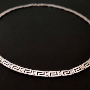 May include: A silver necklace with a Greek key pattern. The necklace is made of a thick, flat chain.