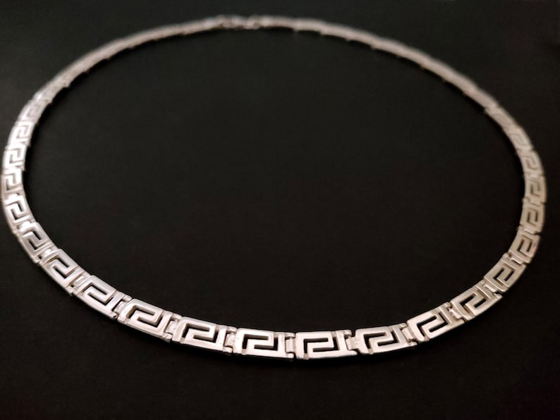 May include: A silver necklace with a Greek key pattern. The necklace is made of a thick, flat chain.