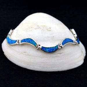 May include: A silver bracelet with a wave-like design, featuring vibrant blue opal inlays. The bracelet is displayed on a white seashell against a black background, showcasing its elegant and eye-catching design.