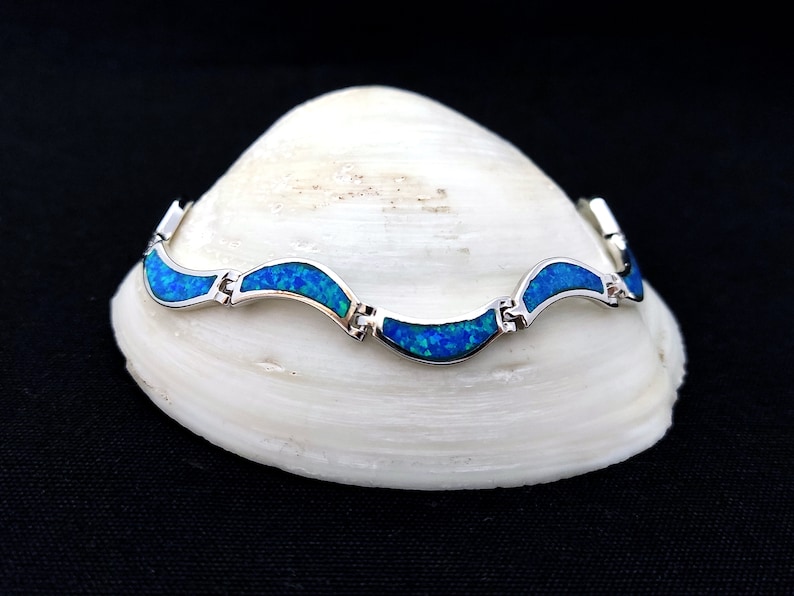 May include: A silver bracelet with a wave-like design, featuring vibrant blue opal inlays. The bracelet is displayed on a white seashell against a black background, showcasing its elegant and eye-catching design.