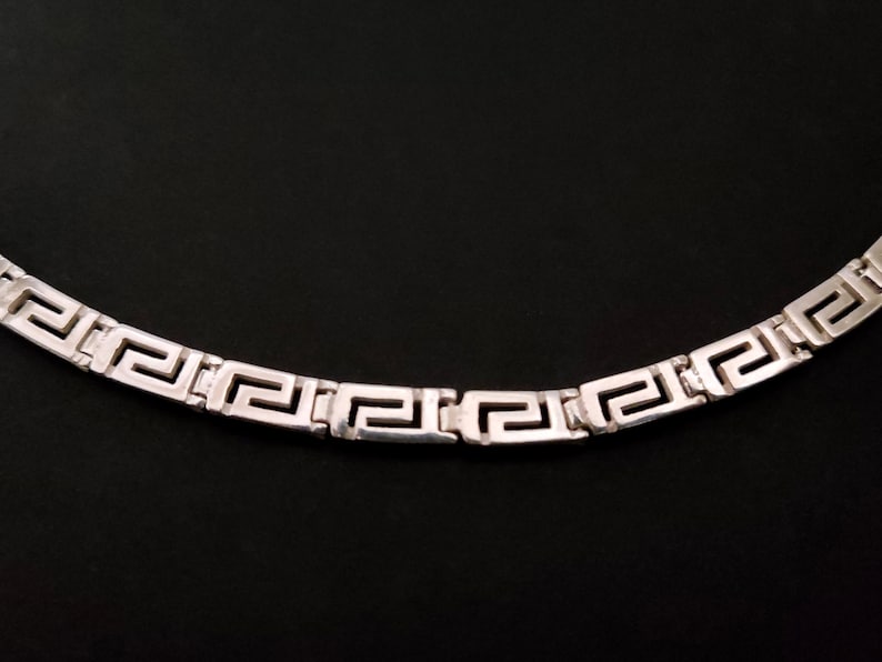 May include: A silver chain necklace with a Greek key pattern.